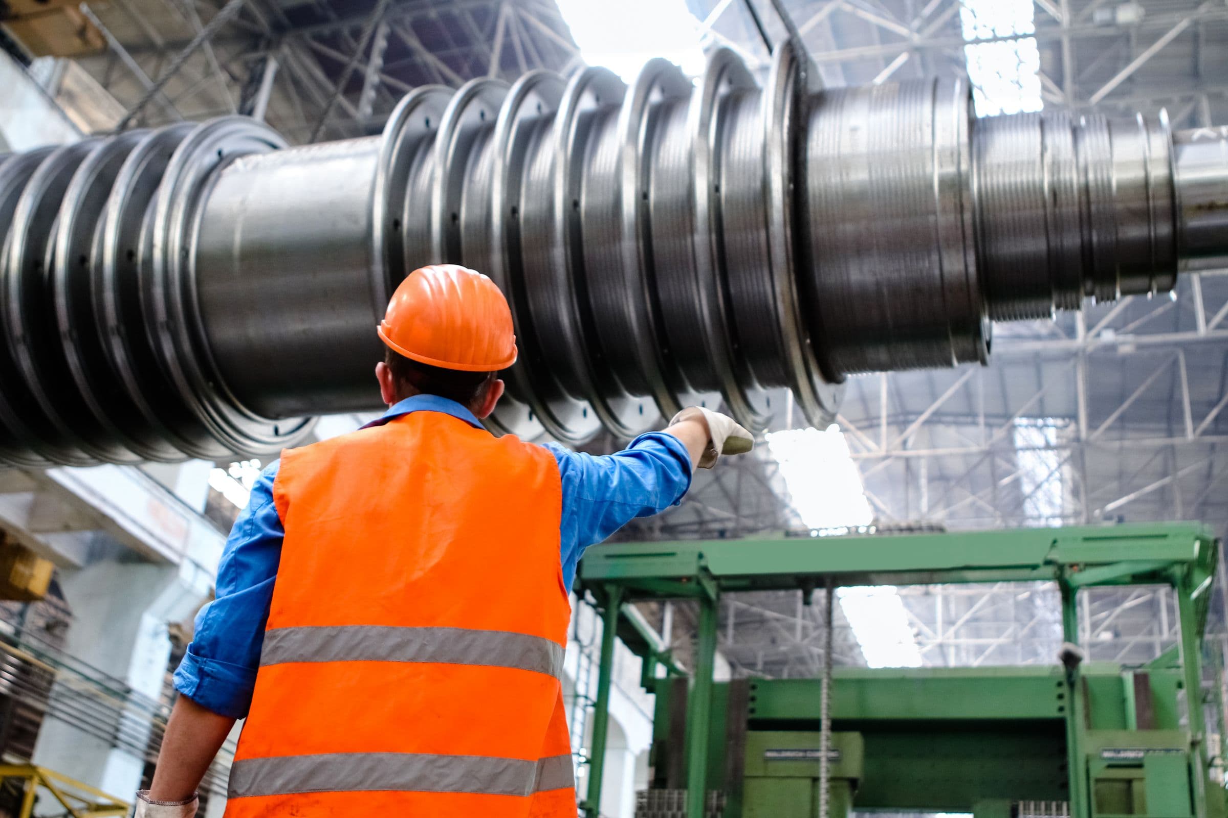 A worker in hard hat and high-visibility vest inspecting a steam turbine rotor inside an industrial plant.