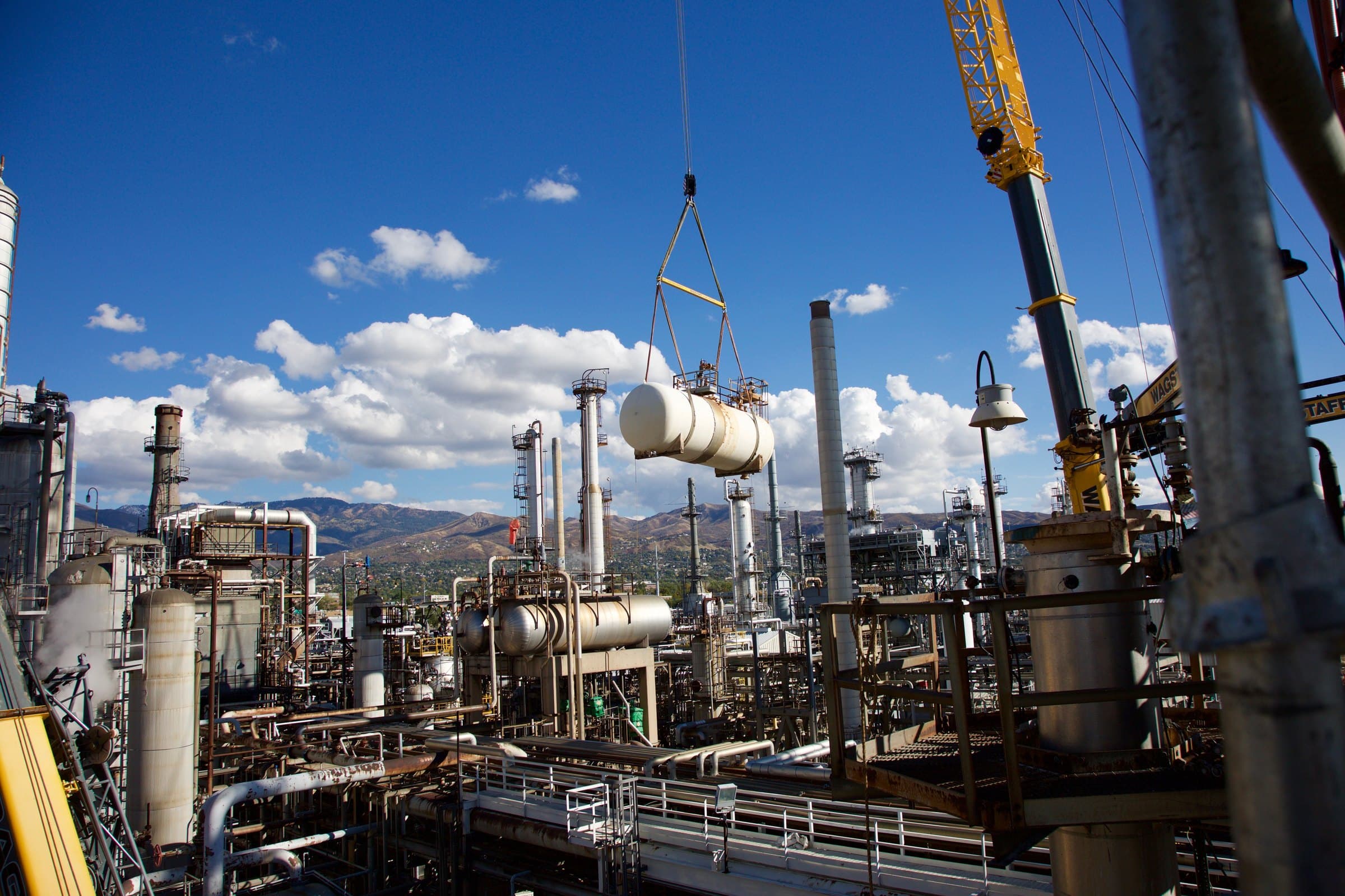 A crane lifting a white horizontal vessel into place at a refinery.
