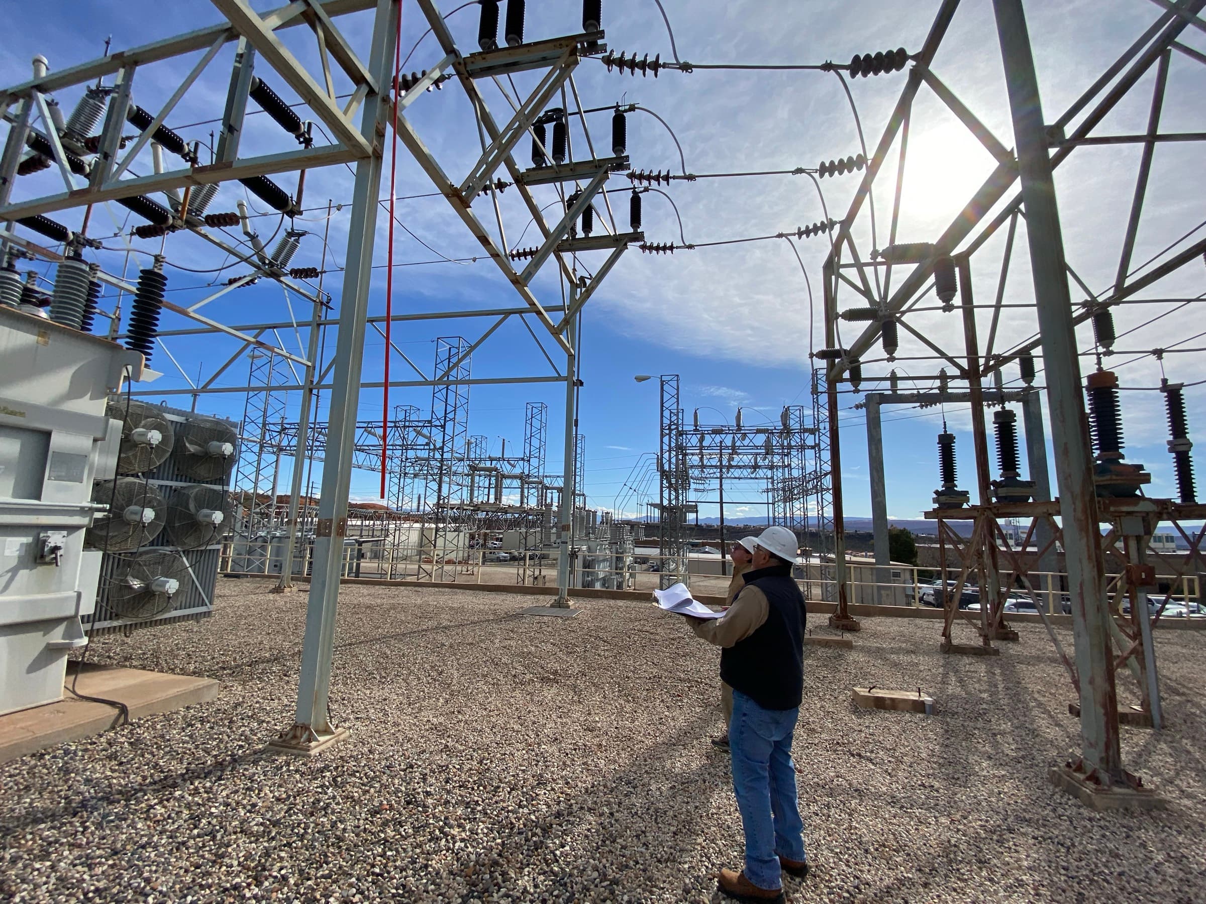 ICPE engineers reviewing drawings inside an energized substation yard, beside a transformer and high-voltage bus structures.